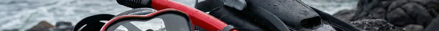 A set of high-quality snorkeling equipment, including a red and black mask, snorkel, and black fins, resting on dark volcanic rocks with a misty coastal cliffside and crashing waves in the background.
